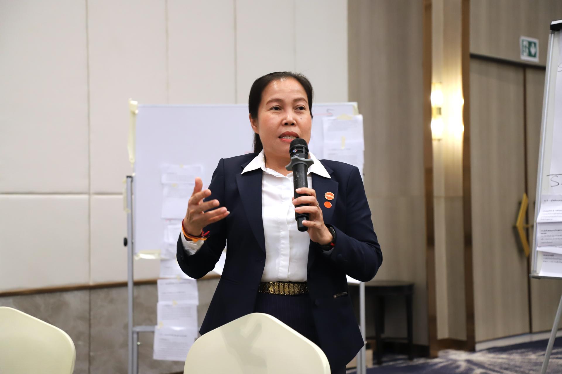 Woman in blazer speaking into a microphone at a conference, with boards in the background.