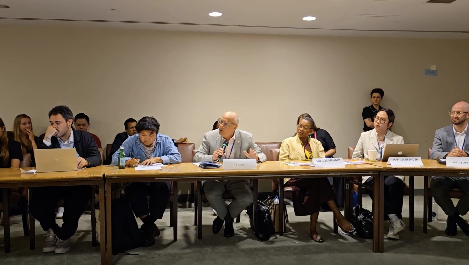Photograph shows a group of people seated at a long conference table in a meeting room, with papers in front.