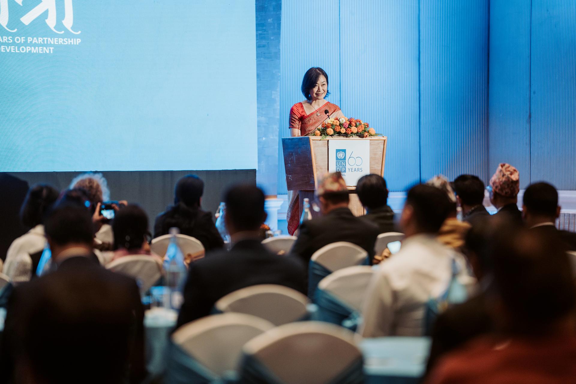 Photo: woman presenting at a podium on a blue stage to conference attendees.
