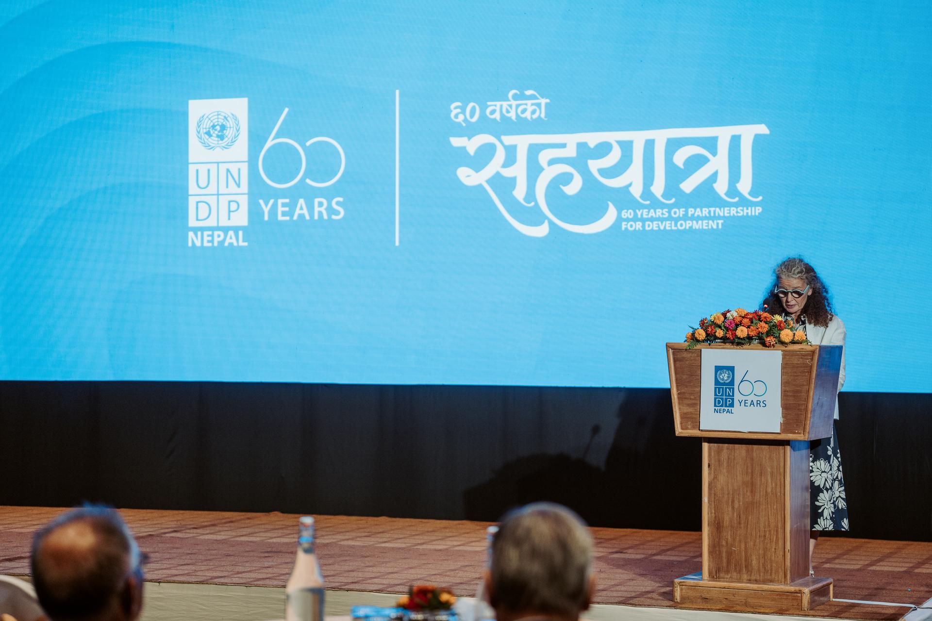 Photograph: woman speaks at a podium on a bright blue stage with UNDP Nepal 60th logo.