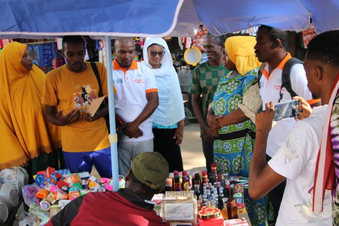 People gather around a market stall under a blue canopy, colorful items on display.
