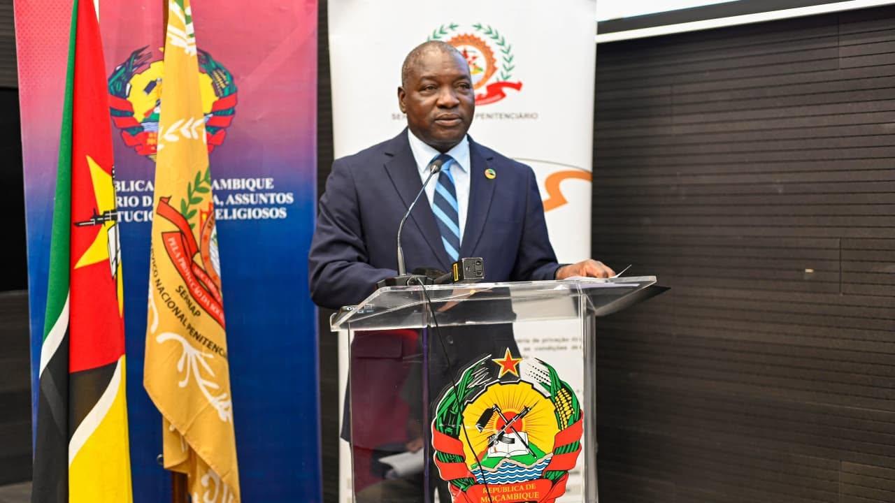A man in a dark suit and blue tie speaks at a podium with a crest; flags and banner behind him.