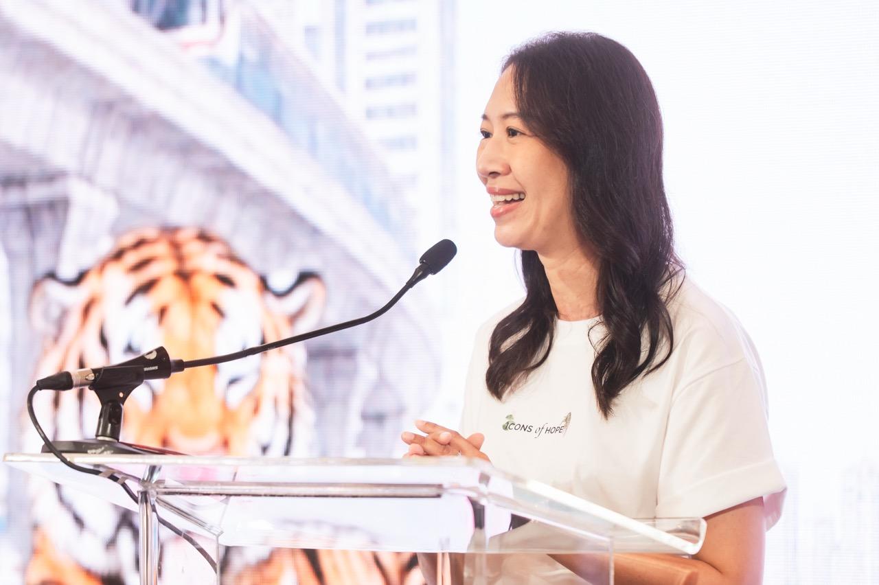 Smiling woman in a white blouse speaks at a clear podium; tiger statue visible behind.