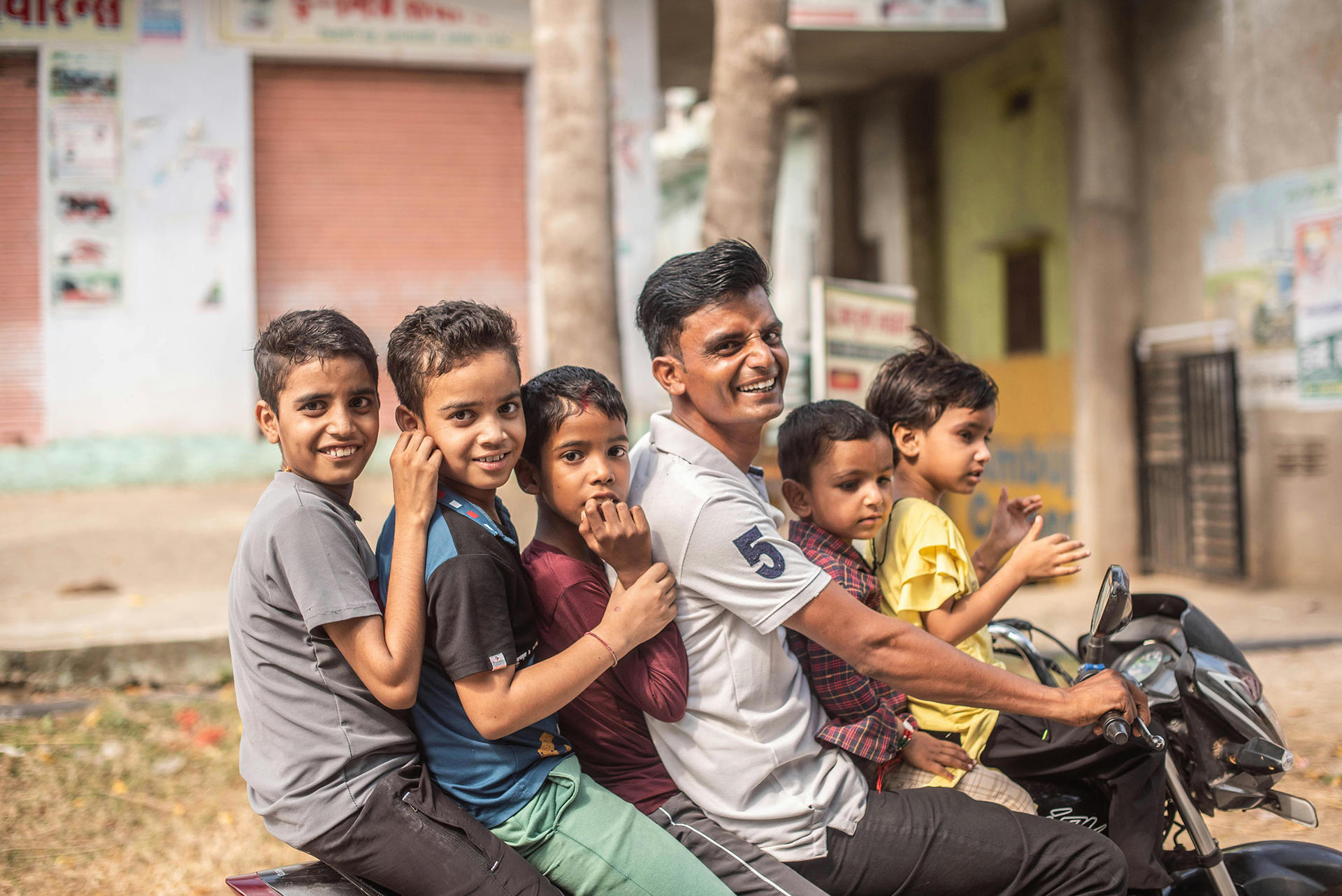 Several kids sit in a line on a single motorcycle on a sunny residential street.
