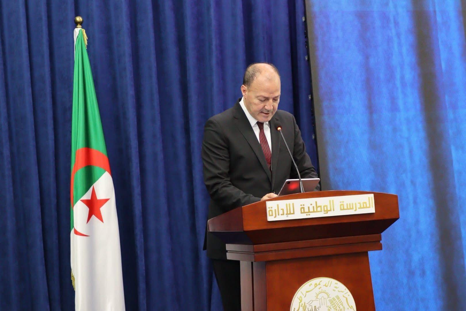 Man at podium giving a speech on a blue stage with the Algerian flag beside him.