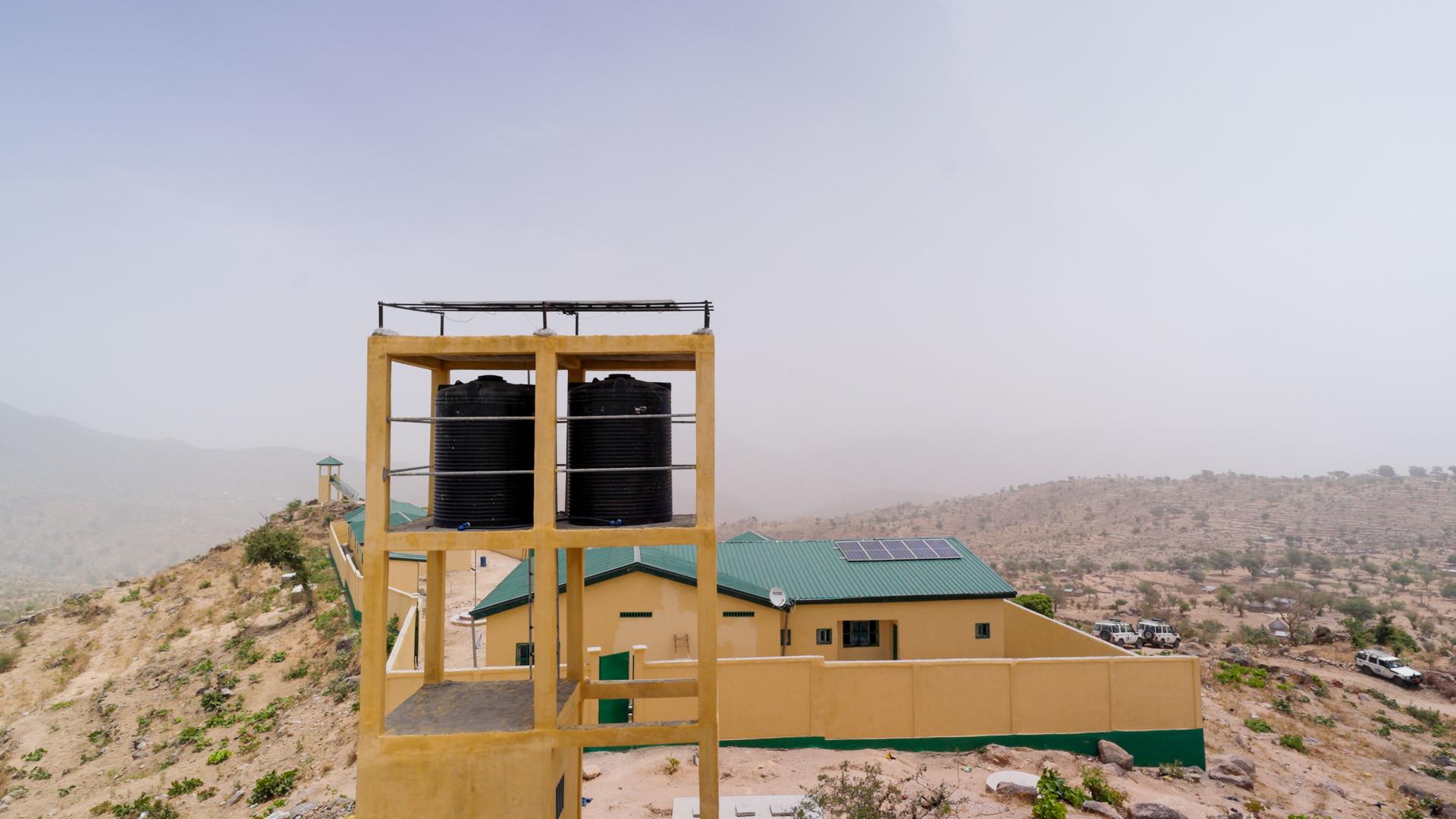 Wooden lookout tower atop a rocky desert hill beside a beige building under a blue sky.