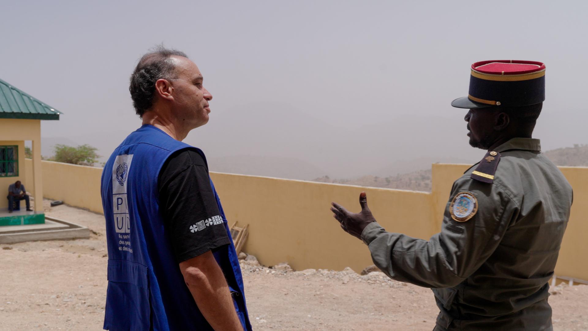 Desert outpost scene with a man in a blue vest speaking to a uniformed officer.