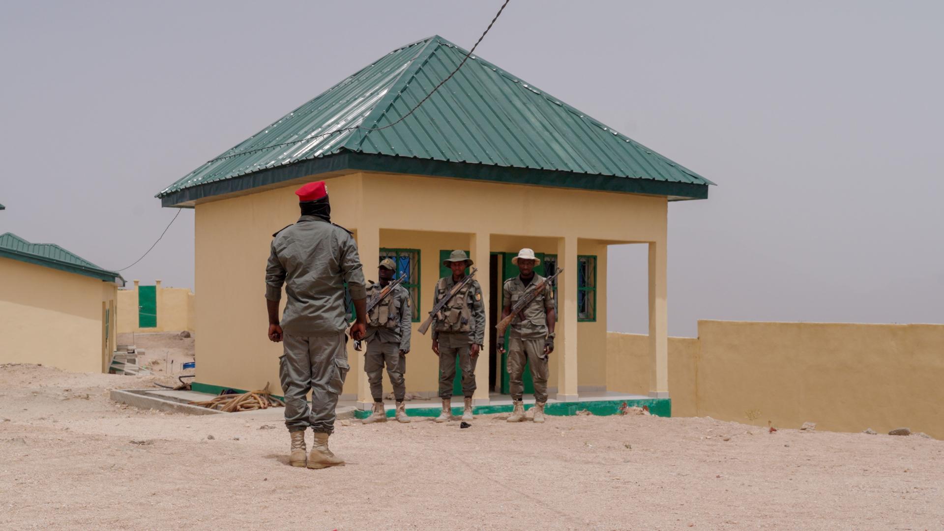 Soldiers in camouflage stand near a small yellow building with a green roof in a dusty yard.