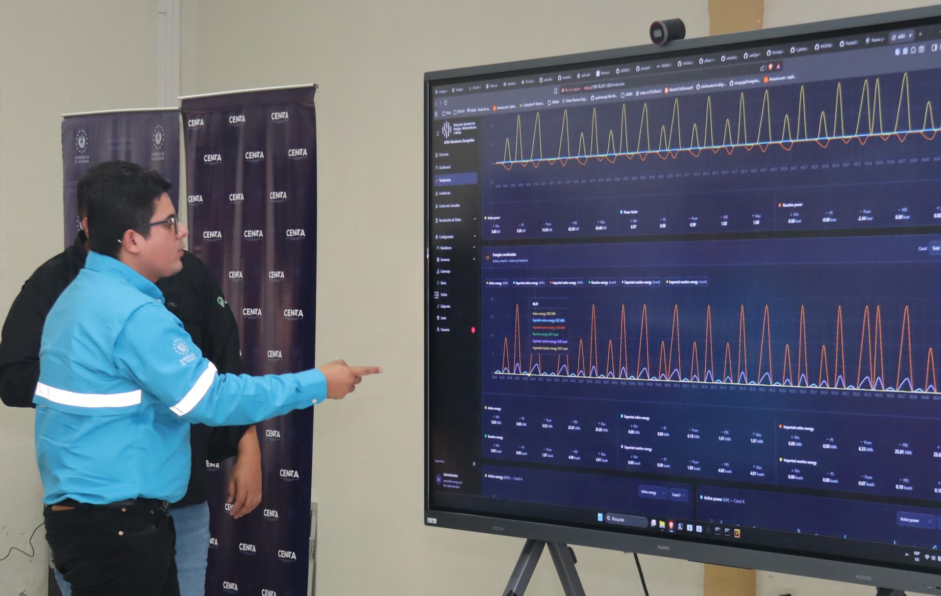 Photo: Two men in blue jackets point at a large screen displaying graphs and data.