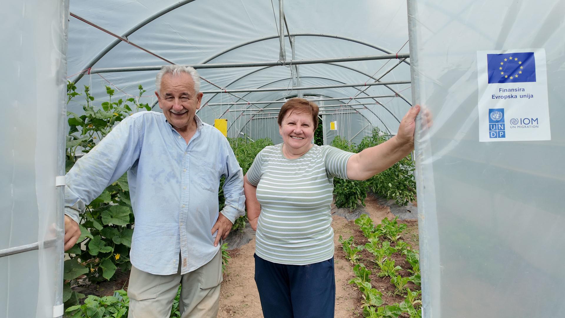 Two people smile at a greenhouse entrance; man in blue shirt, woman in green top.