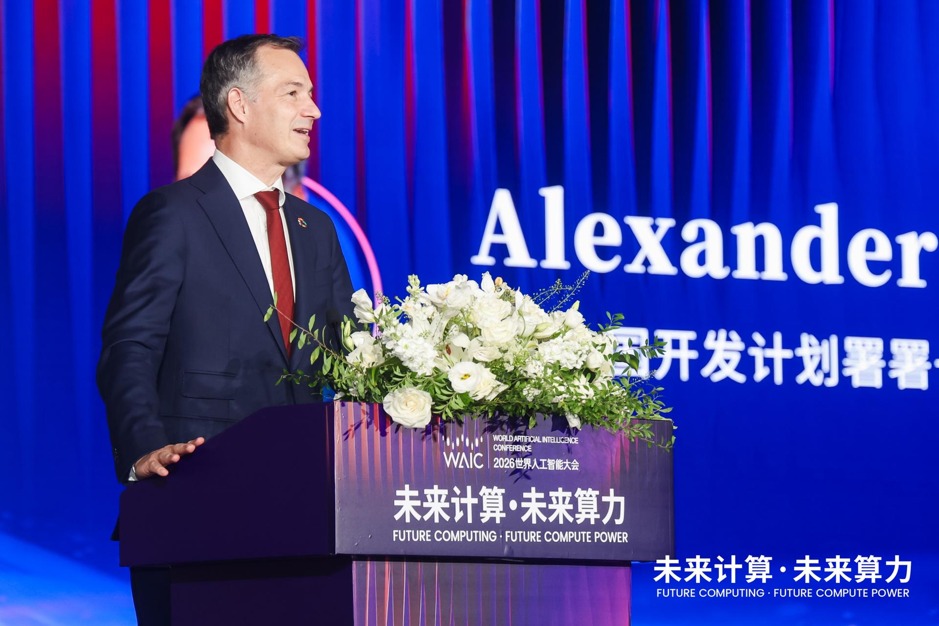 Photo of a man in a dark suit at a podium with white flowers, blue backdrop displaying Alexander.