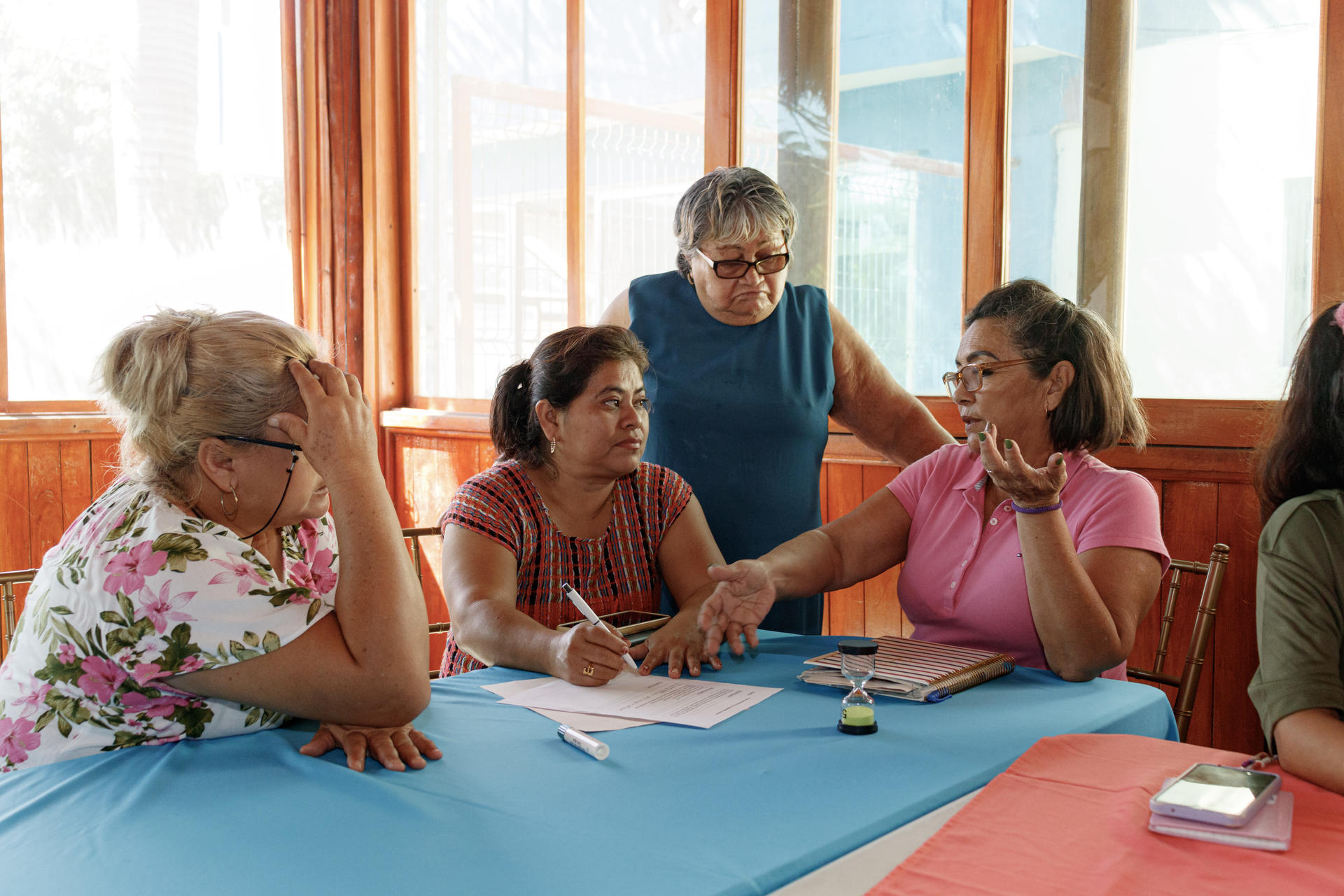 Group of women at a craft workshop around a blue mat, by a sunny window.