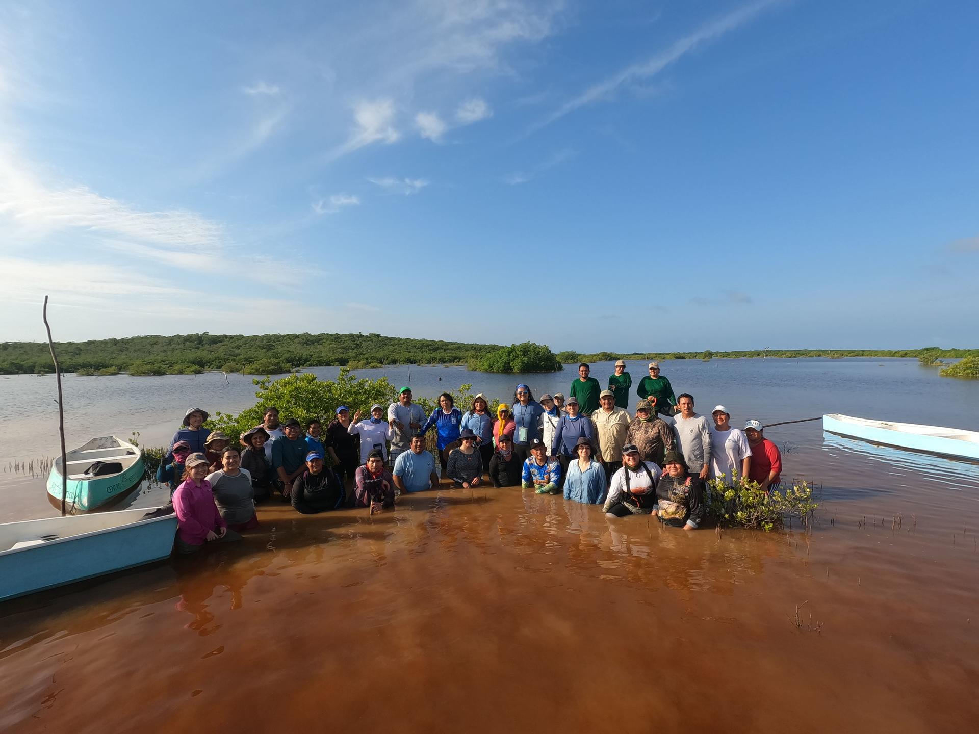 Group of people posing on a muddy riverbank beside a small boat, blue sky and water behind.