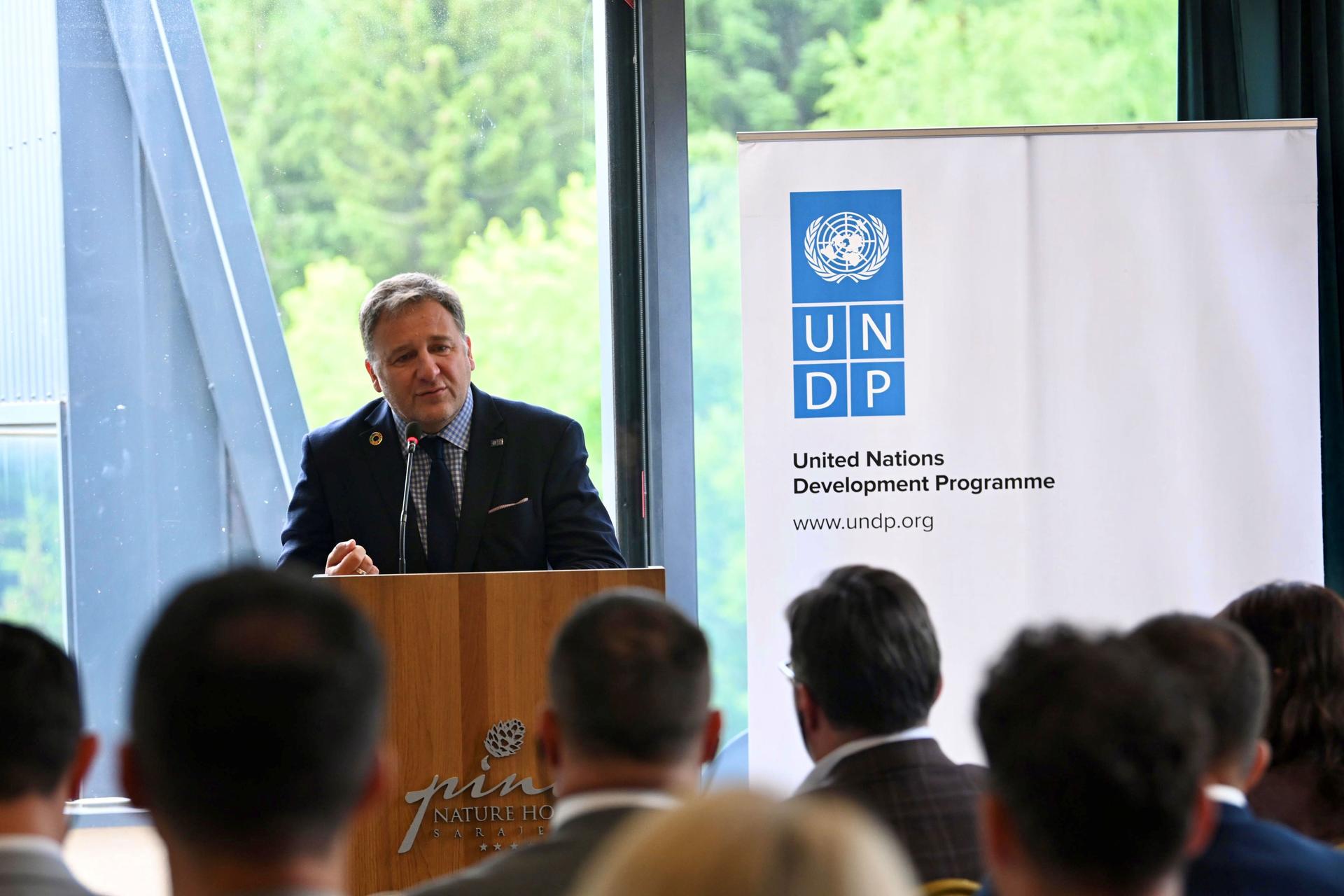 Photograph of a man at a podium addressing an audience, UNDP banner visible.