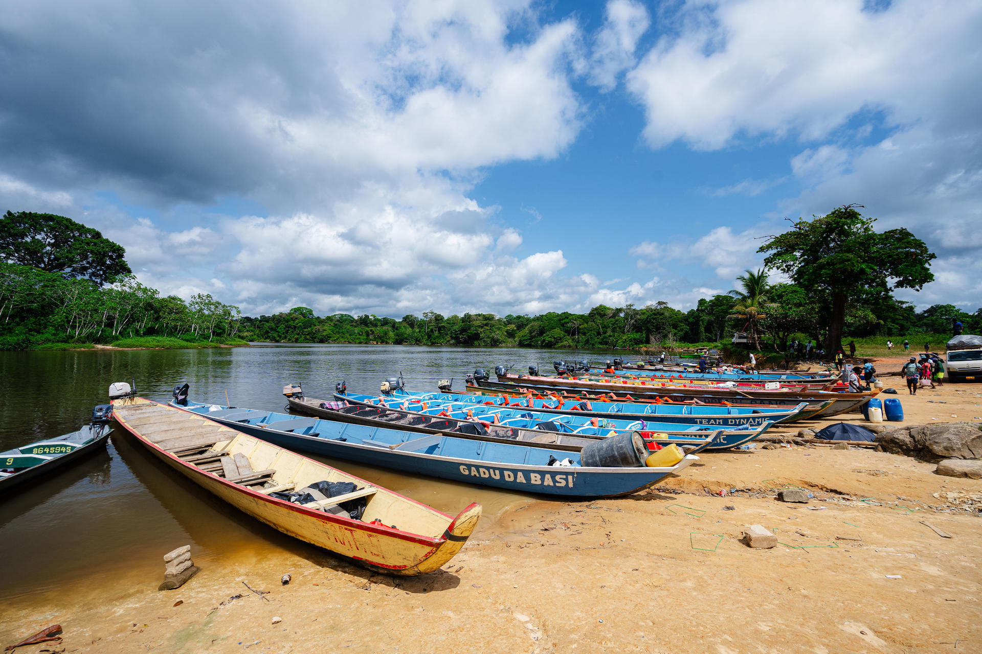Photograph of boats lined on a sandy riverbank with trees and a cloudy sky.