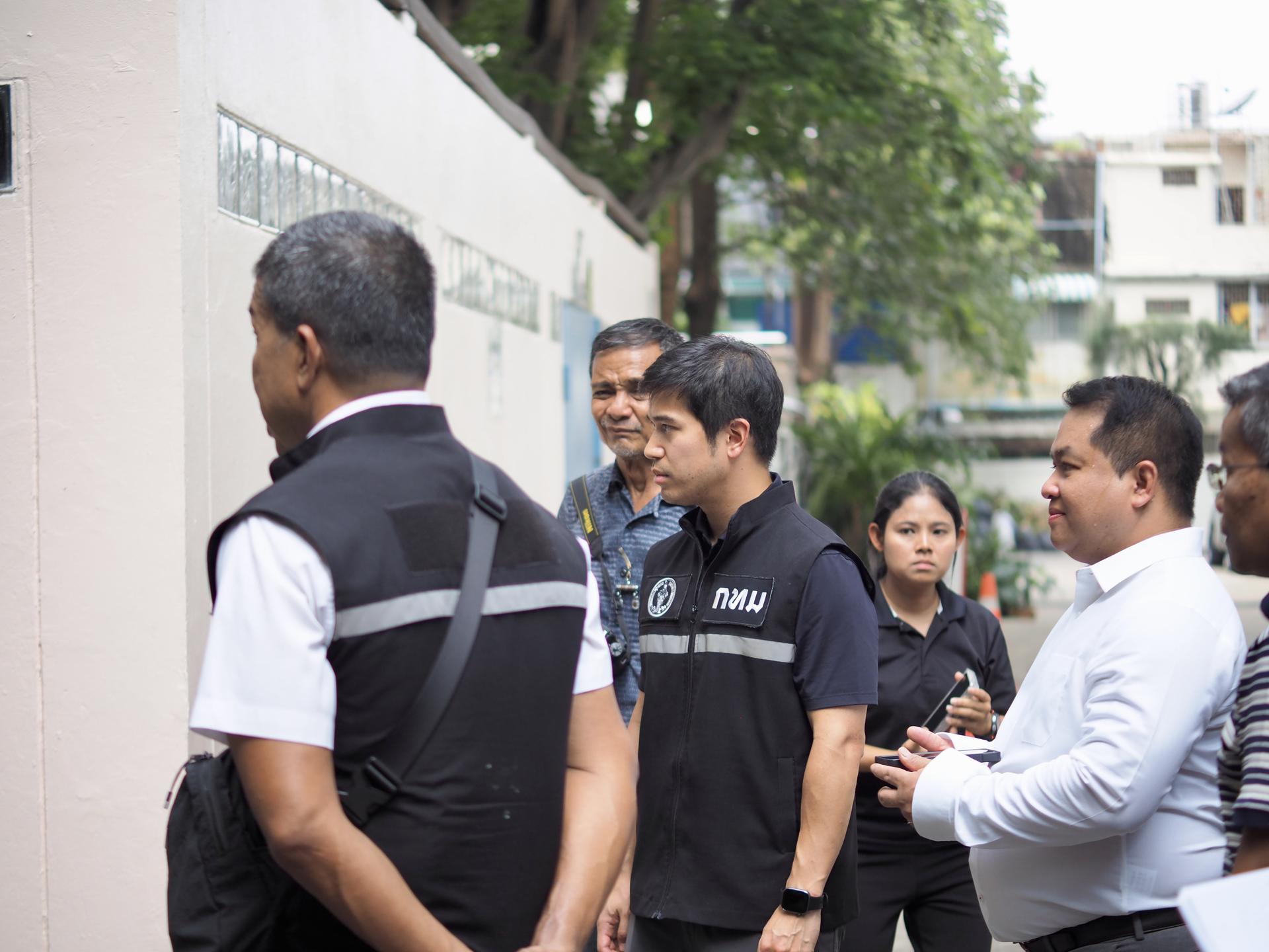 People lined up outside a building along a pink wall, some wearing dark uniforms.