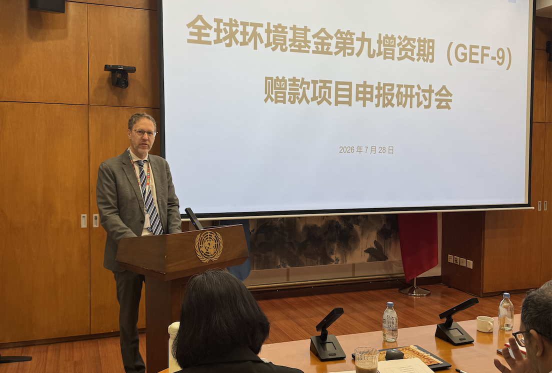 Photograph of a man in a suit at a podium presenting; screen shows Chinese text.