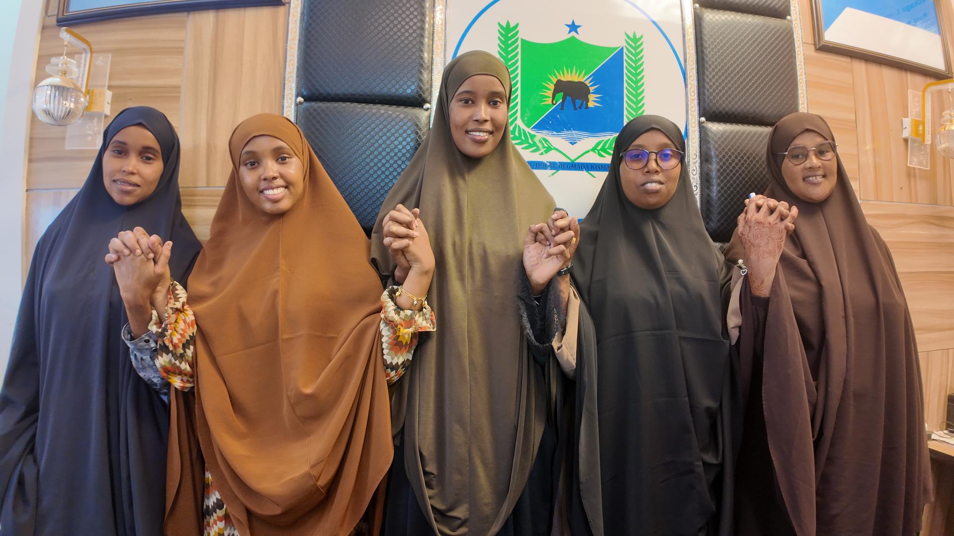 Six women in colorful hijabs and abayas stand in a row on steps in front of a yellow building.