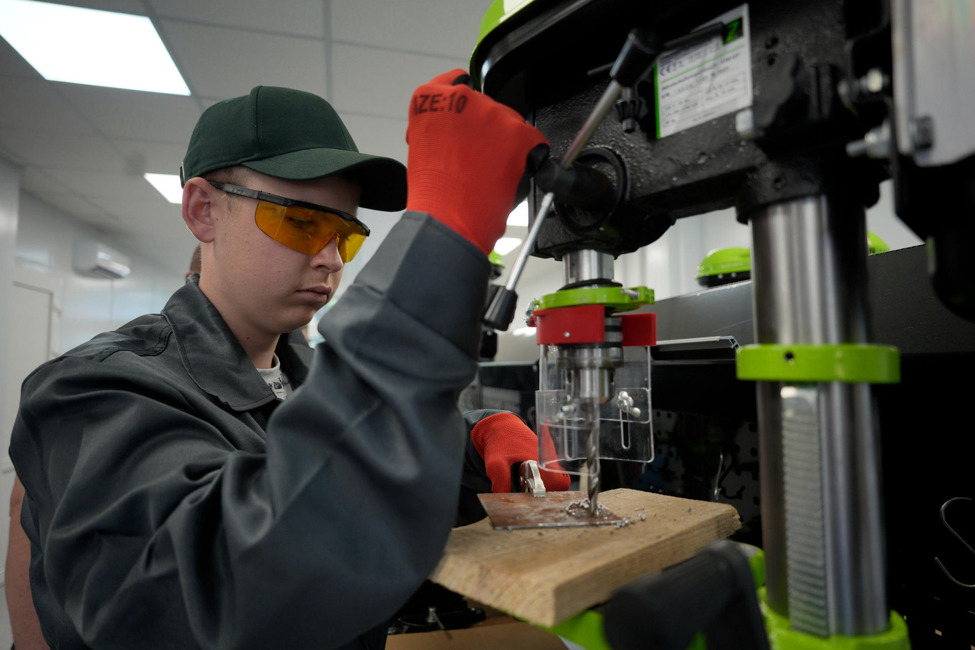 A boy wearing protective goggles, gloves, and a work uniform operates a drilling machine during a practical mechanics lesson in the newly restored modular workshop at Arkhanhelske Vocational Agricultural Lyceum in Kherson Oblast, supported by UNDP and its partners.