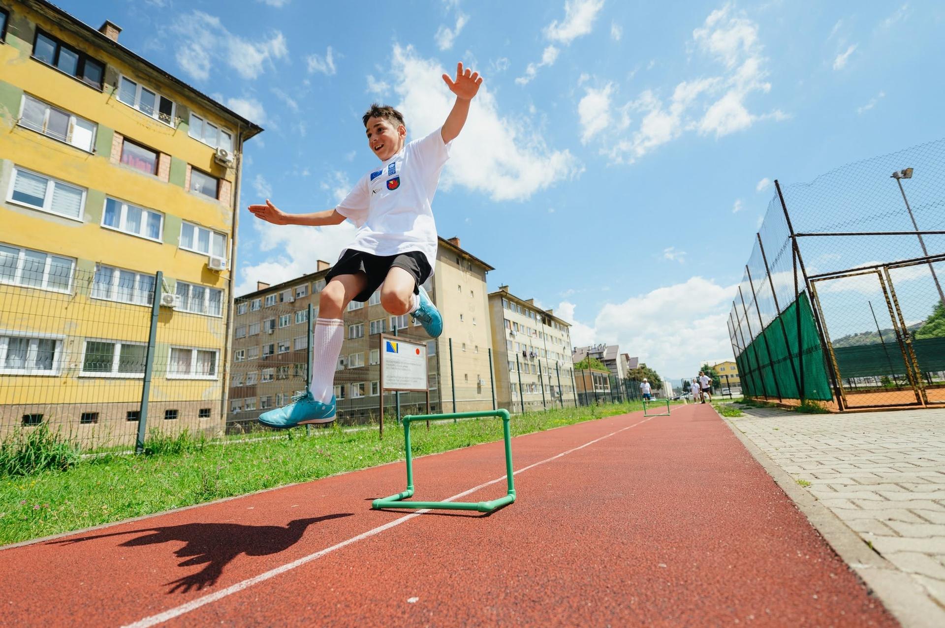 Young athlete leaping over a hurdle on a red track with yellow apartment buildings in background