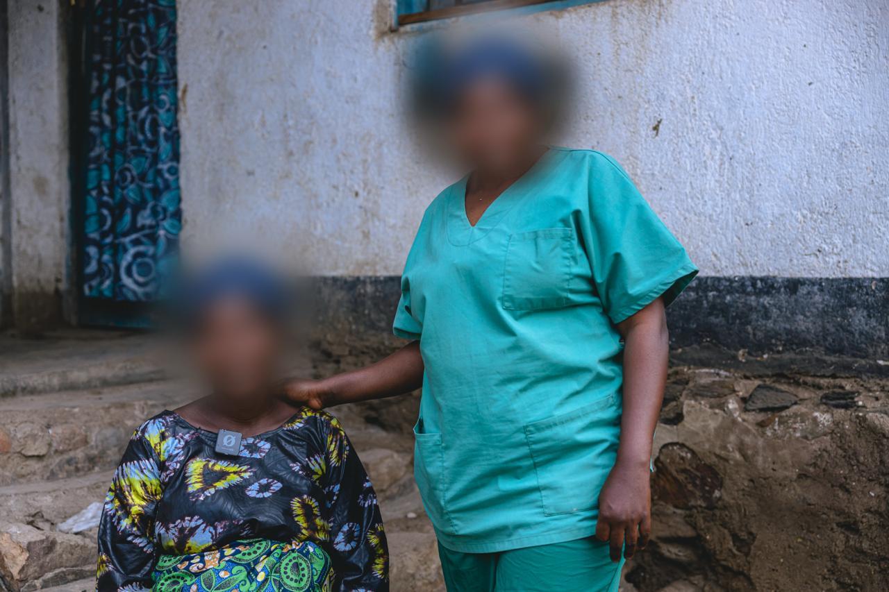 Caregiver in teal scrubs stands beside a seated person in patterned clothing; faces blurred outdoors.