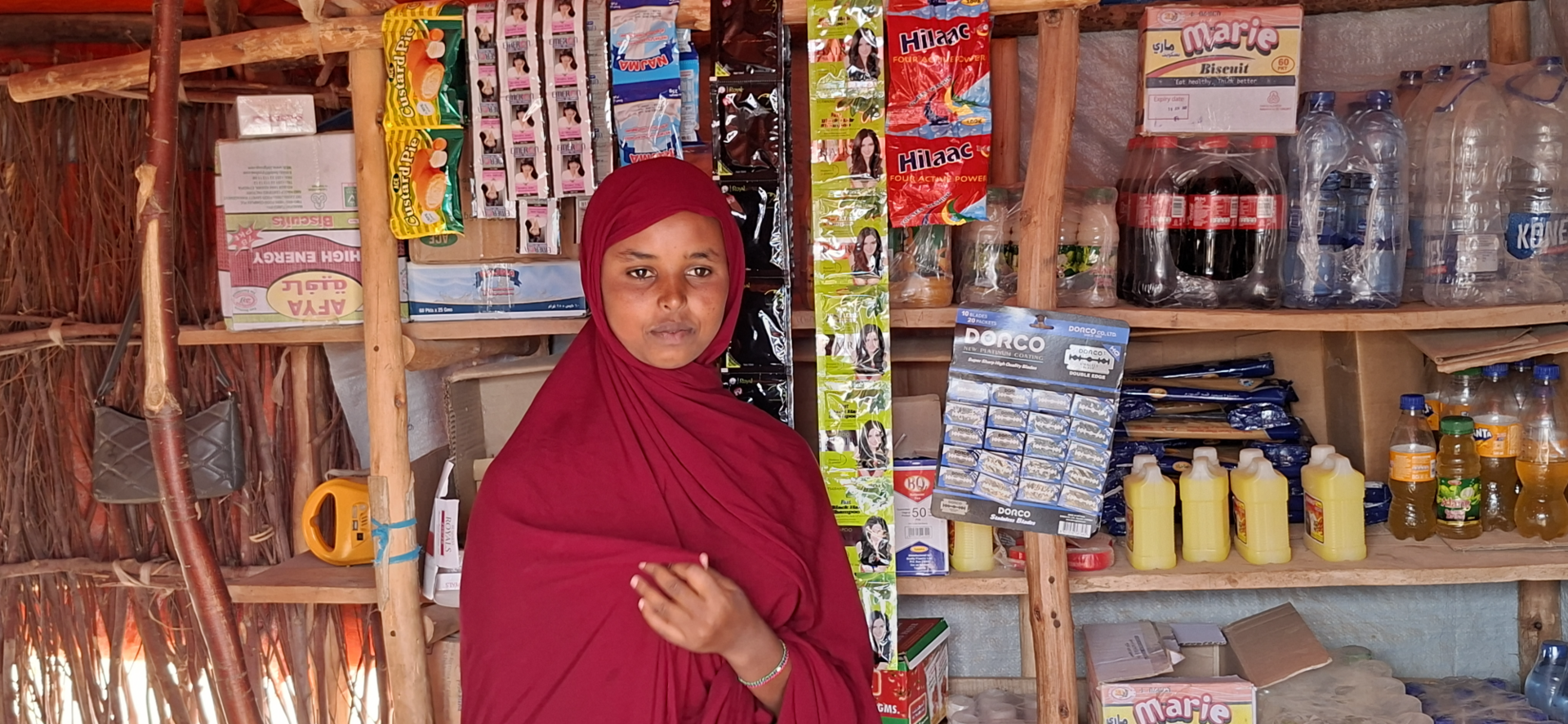 Photograph: woman in red hijab standing in a small market shop with shelves of groceries.