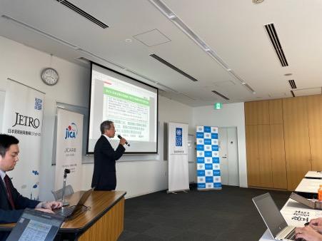 Photo: Presenter delivering a slide presentation to a small audience with laptops in a conference room.
