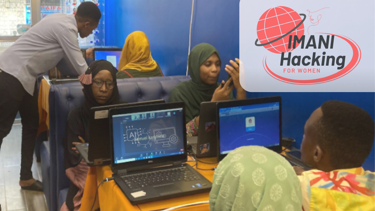 Group of students around laptops in a computer lab; a man helps at a table; banner visible in background.