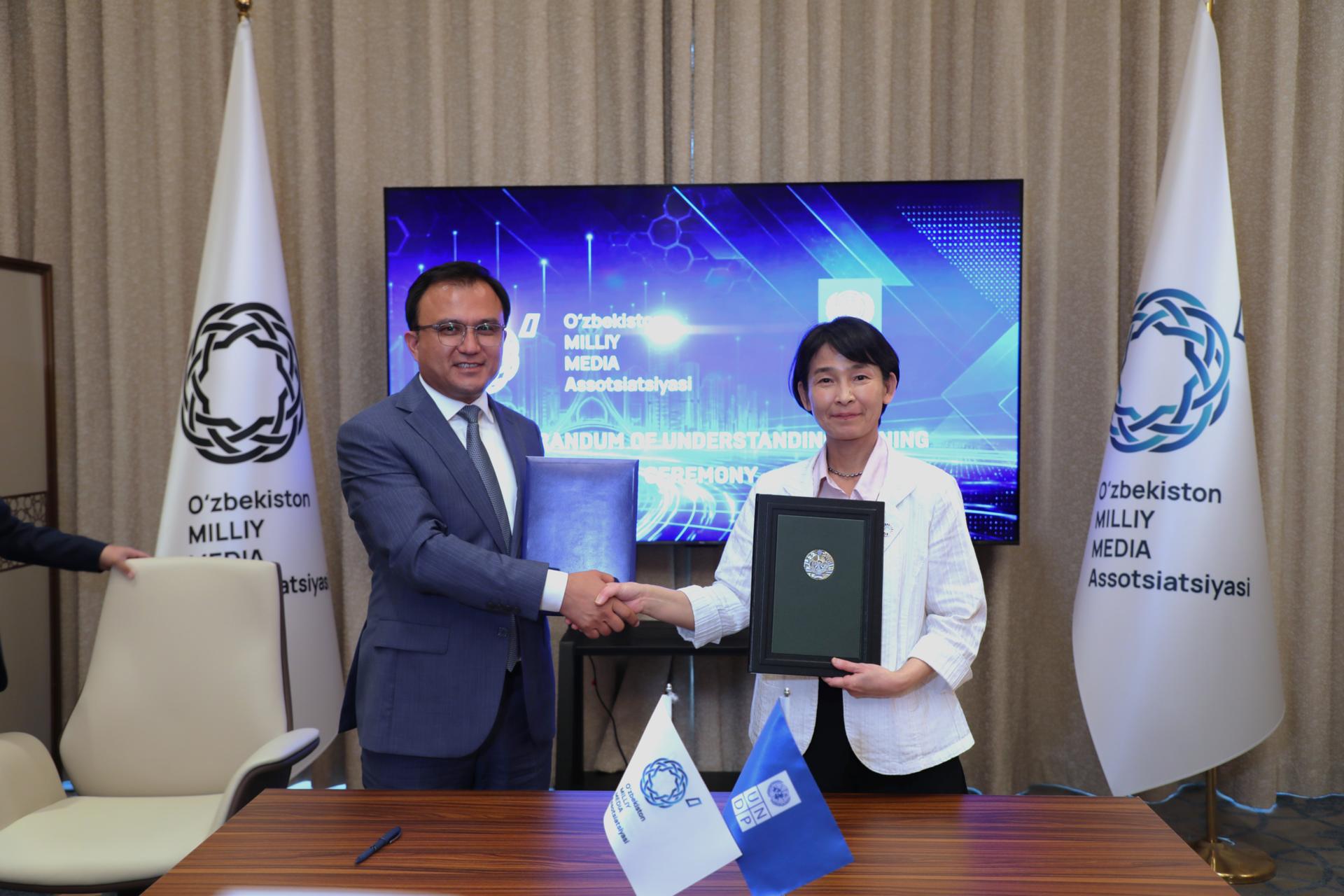 Photograph: two professionals shake hands after signing, flags and a blue banner behind.