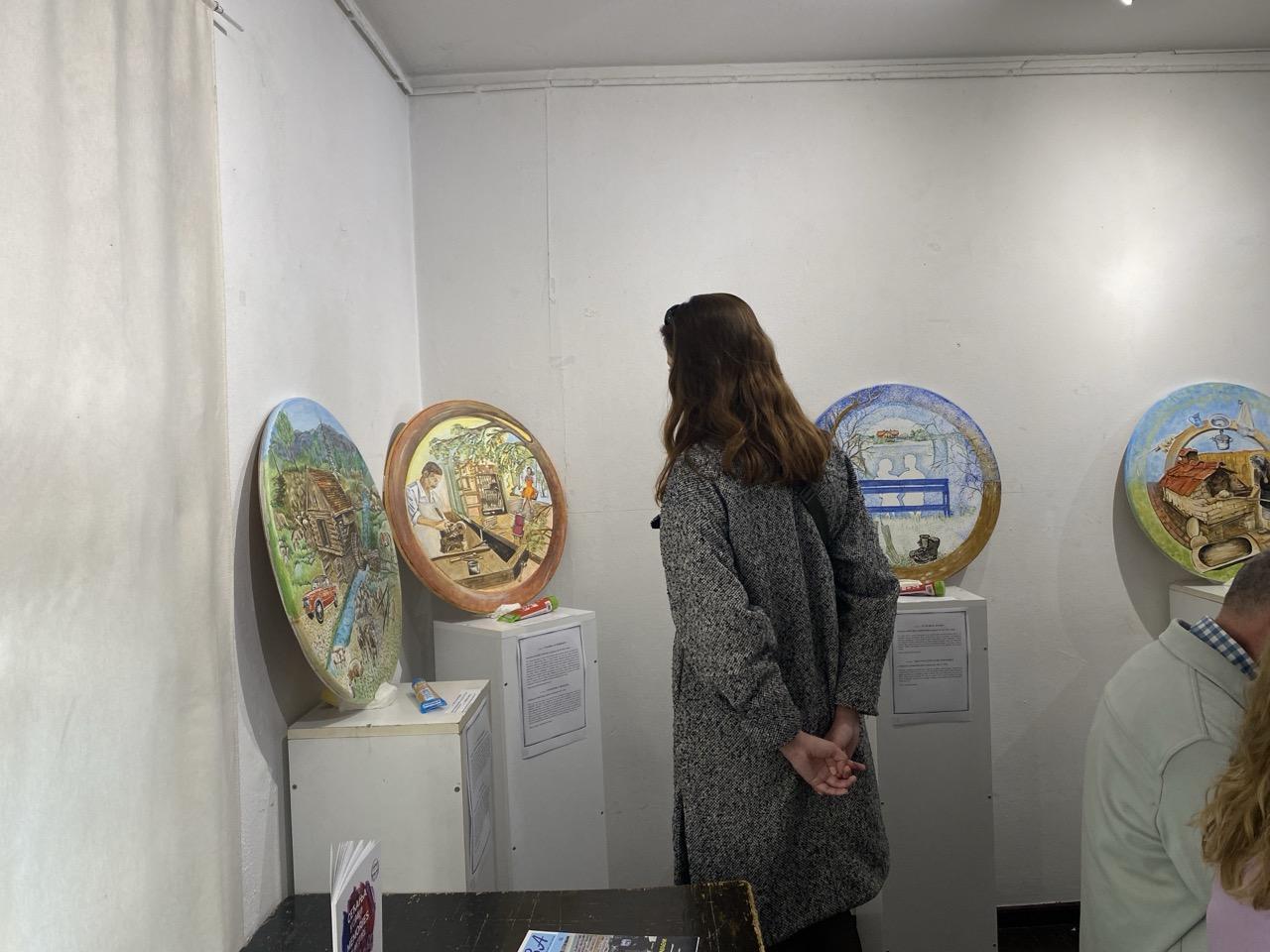 Woman in a gray coat stands in a gallery, viewing circular artworks on the wall.
