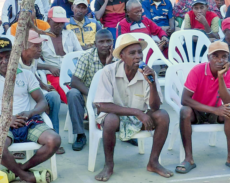 Group of men seated on plastic chairs outdoors, one man speaks into a microphone.
