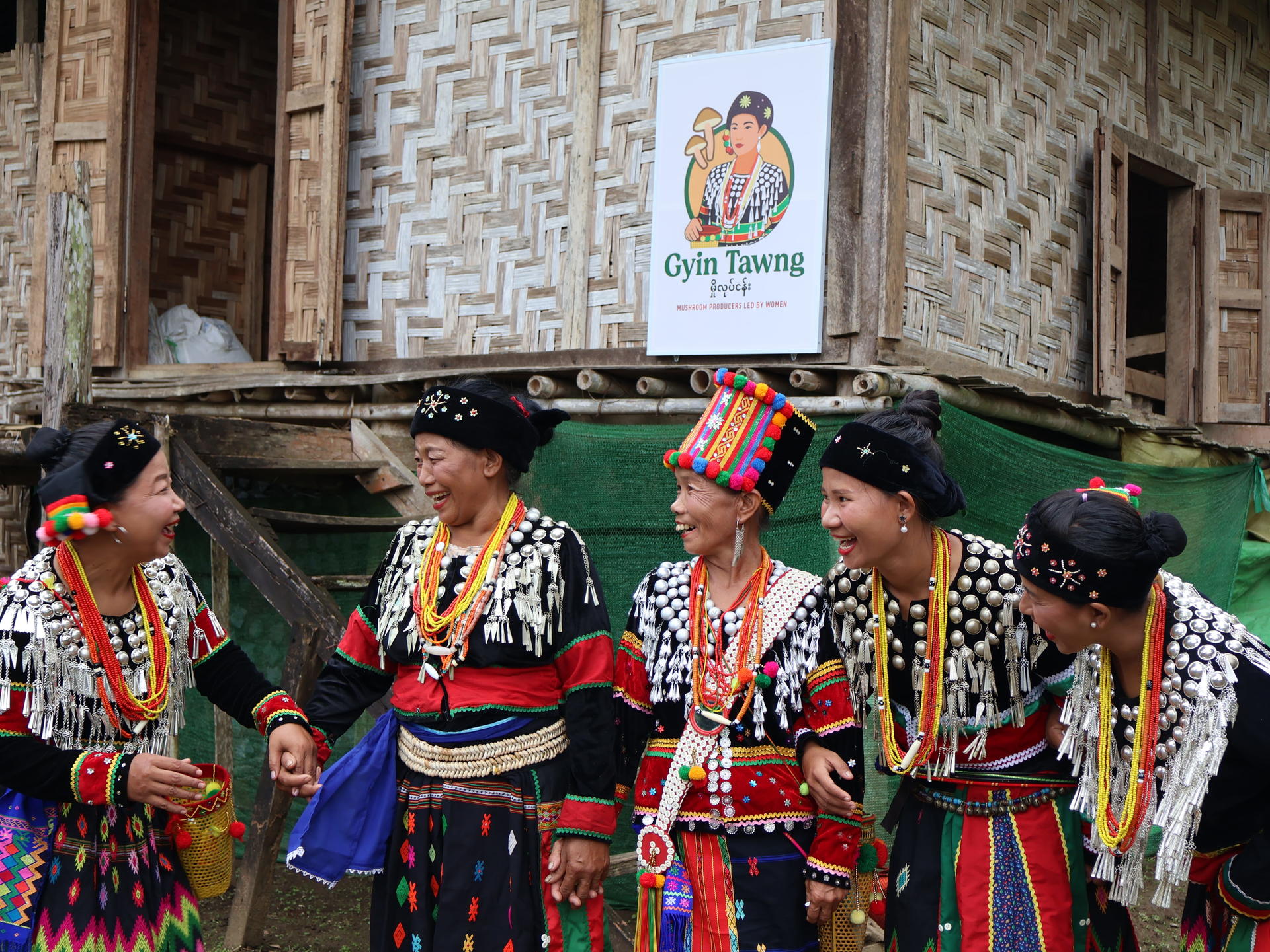 Group in traditional dress with bead necklaces and hats, standing in a woven bamboo doorway.