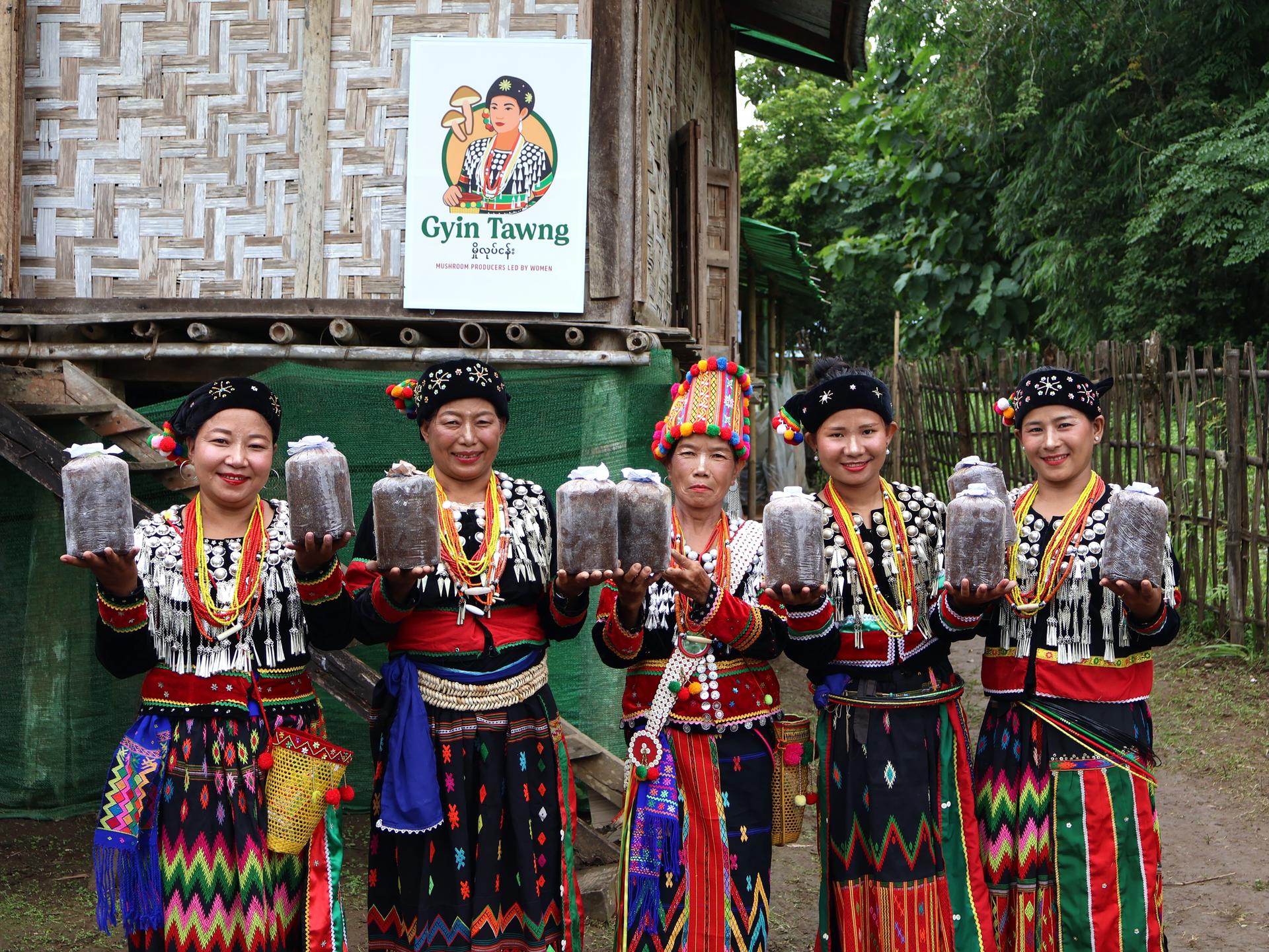 Five people in colorful traditional outfits pose outdoors in front of a wooden building with a sign.