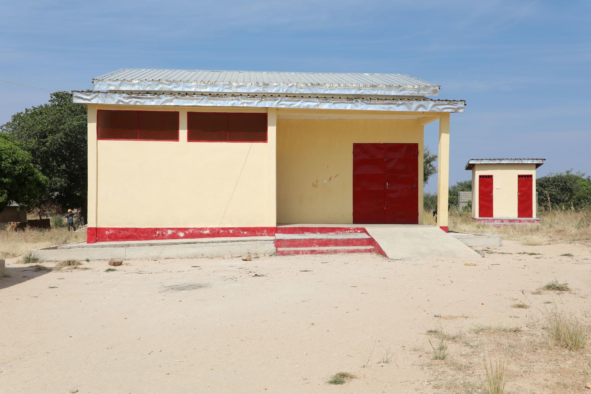 Small beige building with red door and steps in a sandy desert under a blue sky.