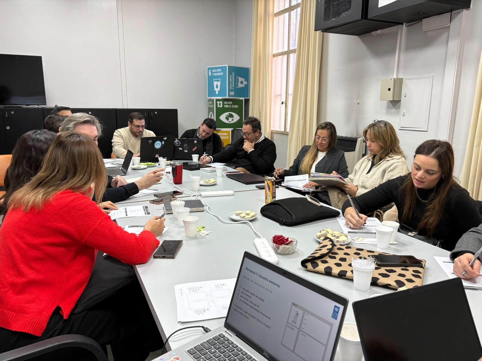 Group of people around a conference table in a meeting room with laptops, papers, and snacks.