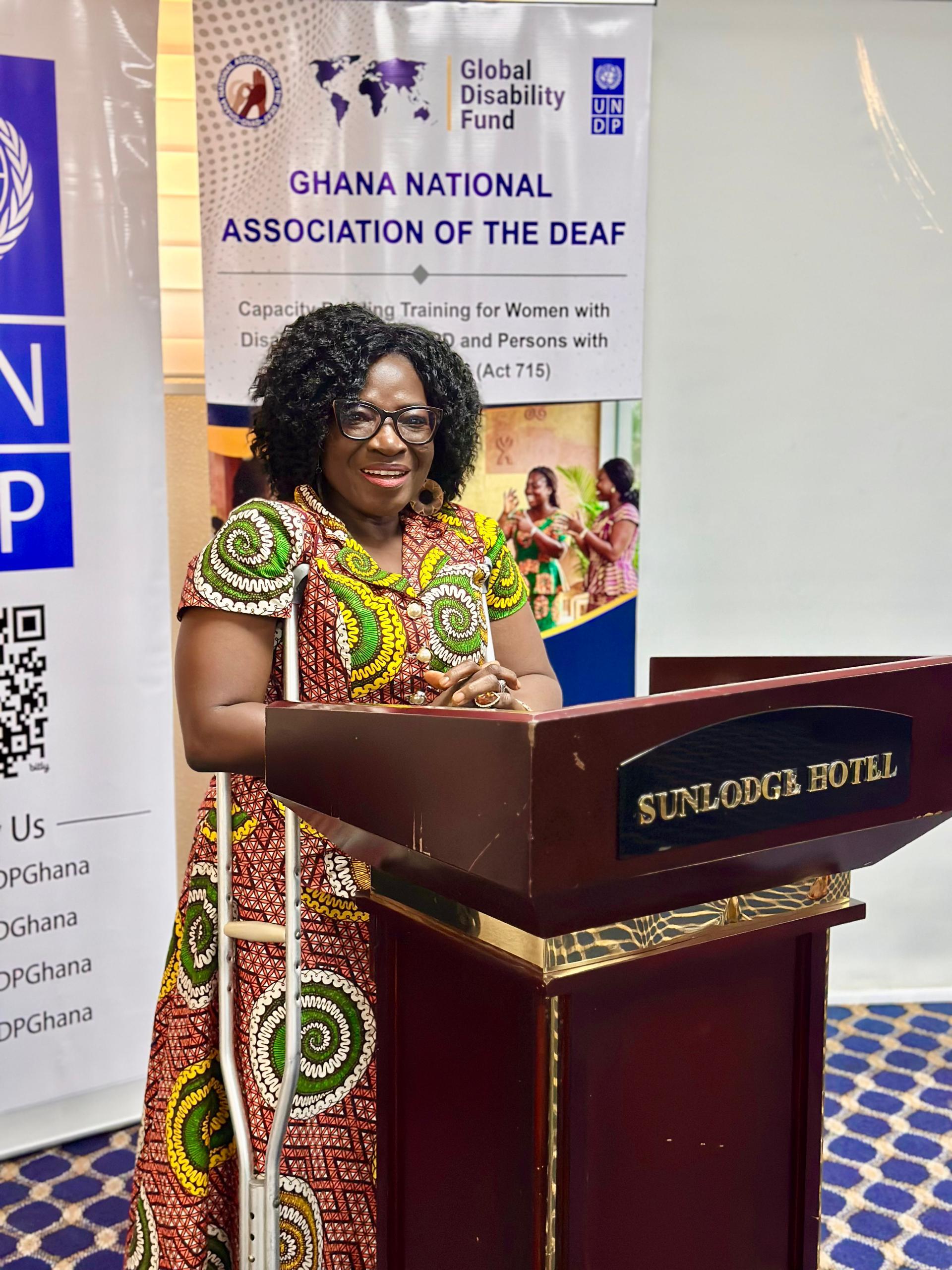 A woman in a colorful patterned dress speaks at a podium during a Ghana National Association of the Deaf event.