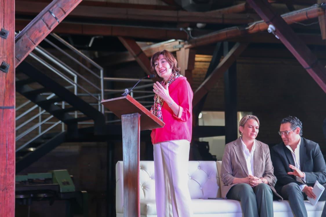 Woman in pink on stage raises arm while two men sit on a couch.
