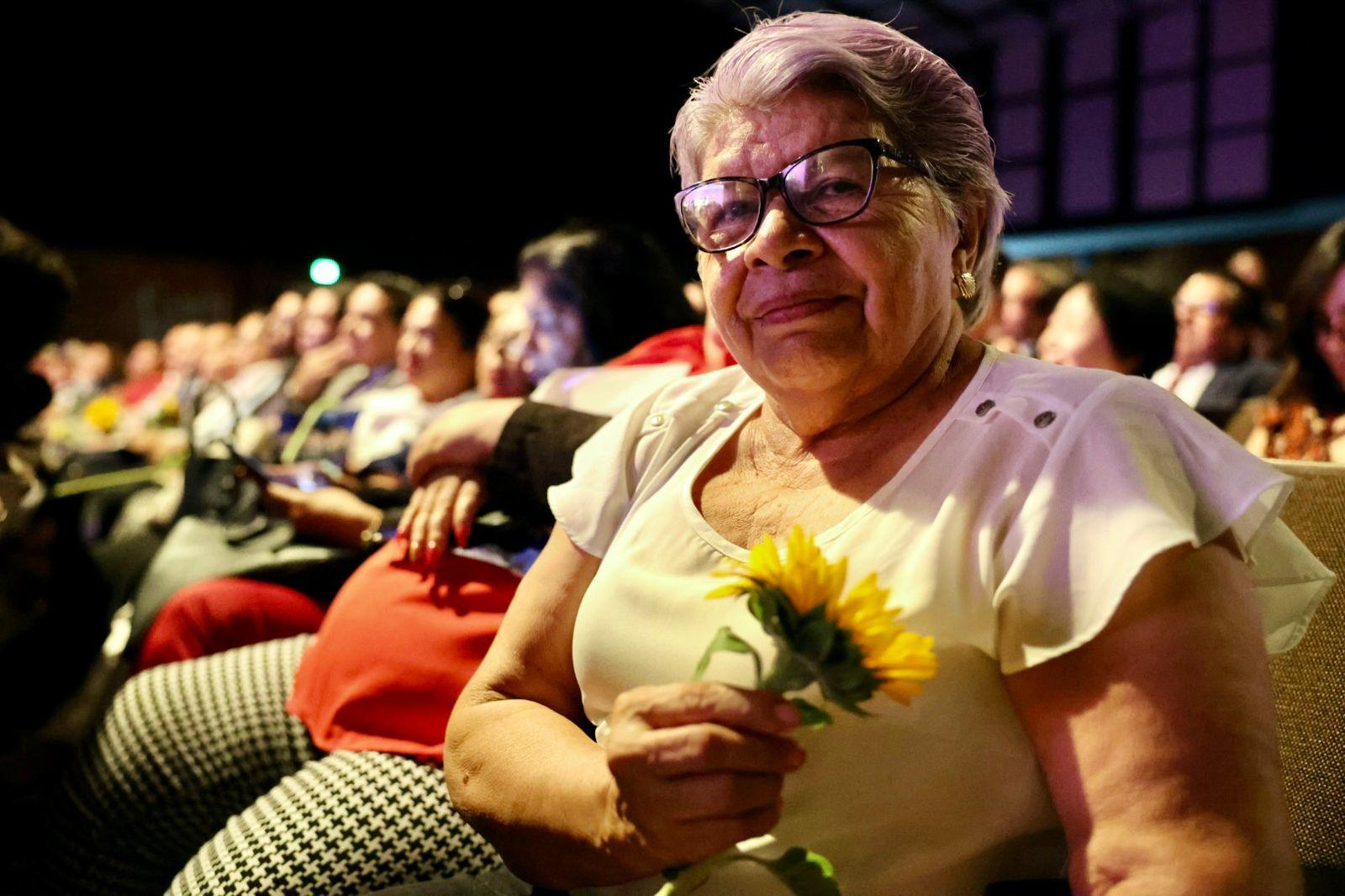 An elderly woman in a white blouse holds a yellow flower while seated in an audience at a theater.