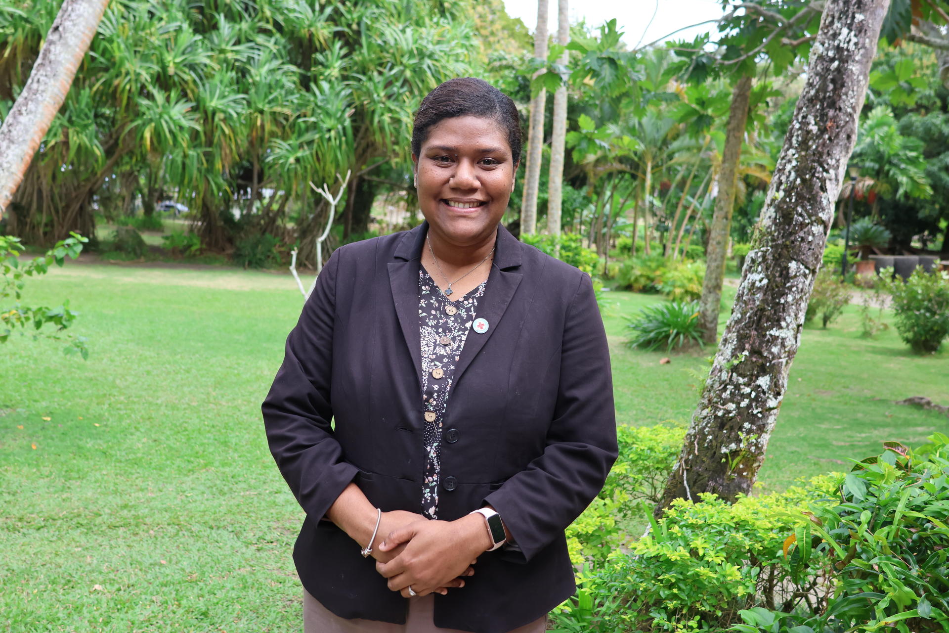 Smiling woman in a dark blazer standing in a lush garden with trees.