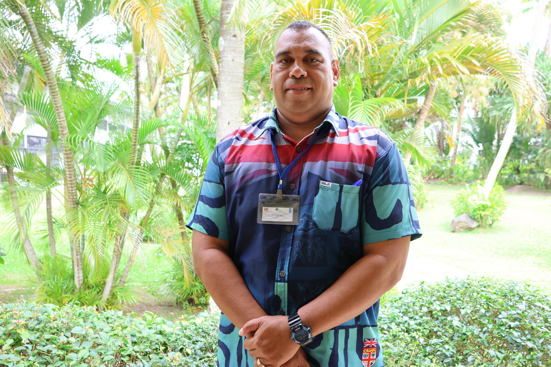 Man in a striped shirt with a lanyard badge standing in a tropical garden.