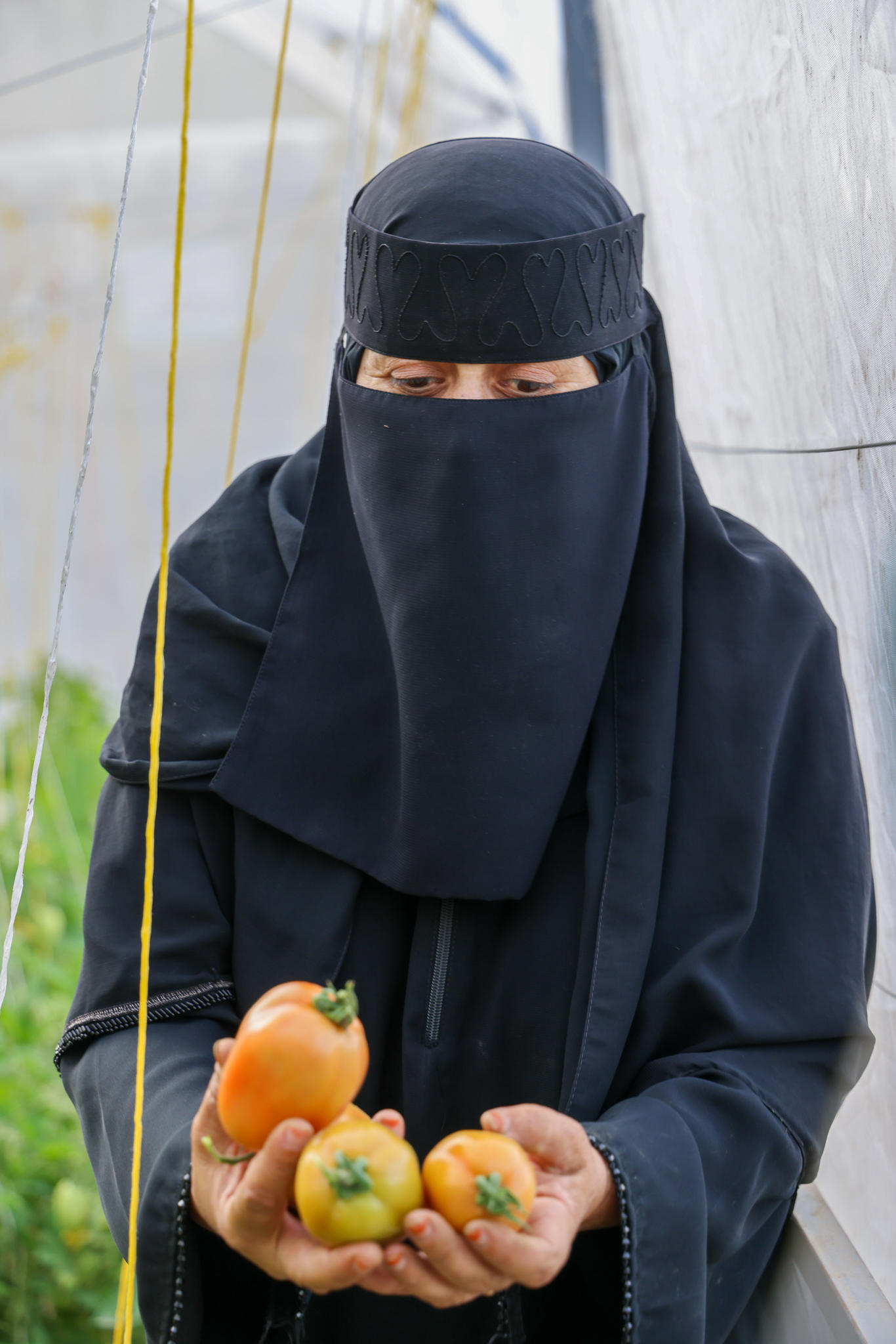 Person in a black niqab and cloak holds several small orange fruits outdoors.