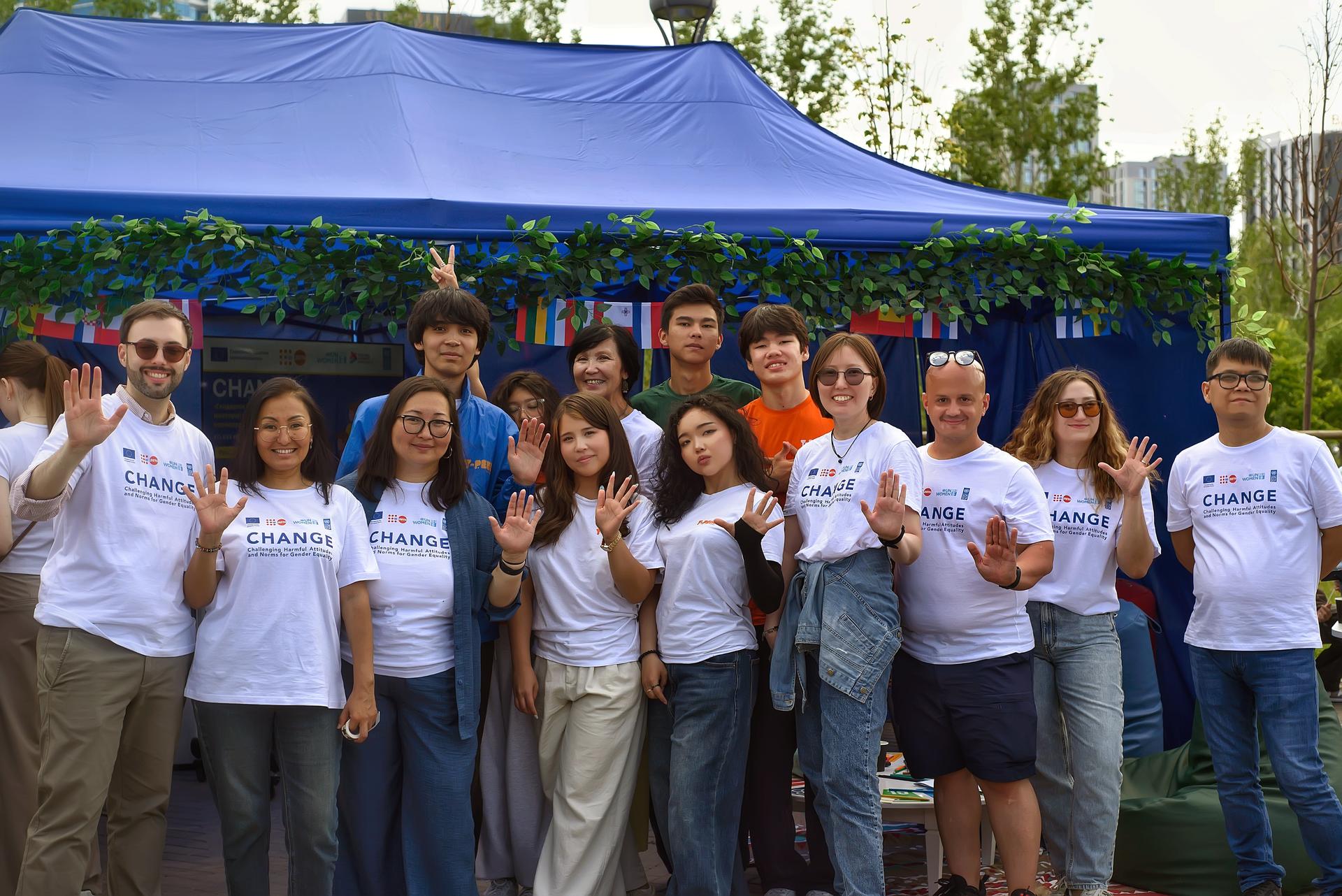 Group of people in white t-shirts posing outdoors under a blue canopy, some giving thumbs up.
