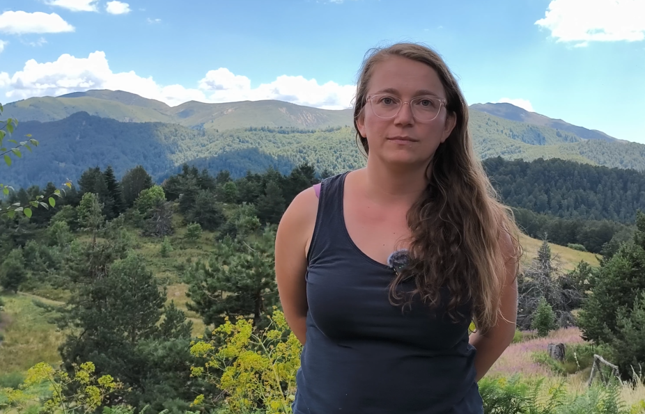 Photograph of a woman with long hair standing outdoors, with hills and forest behind.