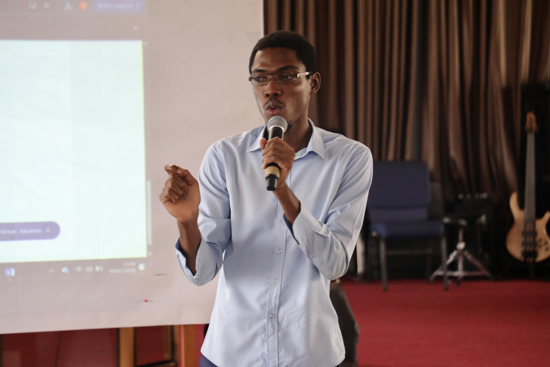 Man in light blue shirt speaks into microphone during presentation, projector screen to the side.
