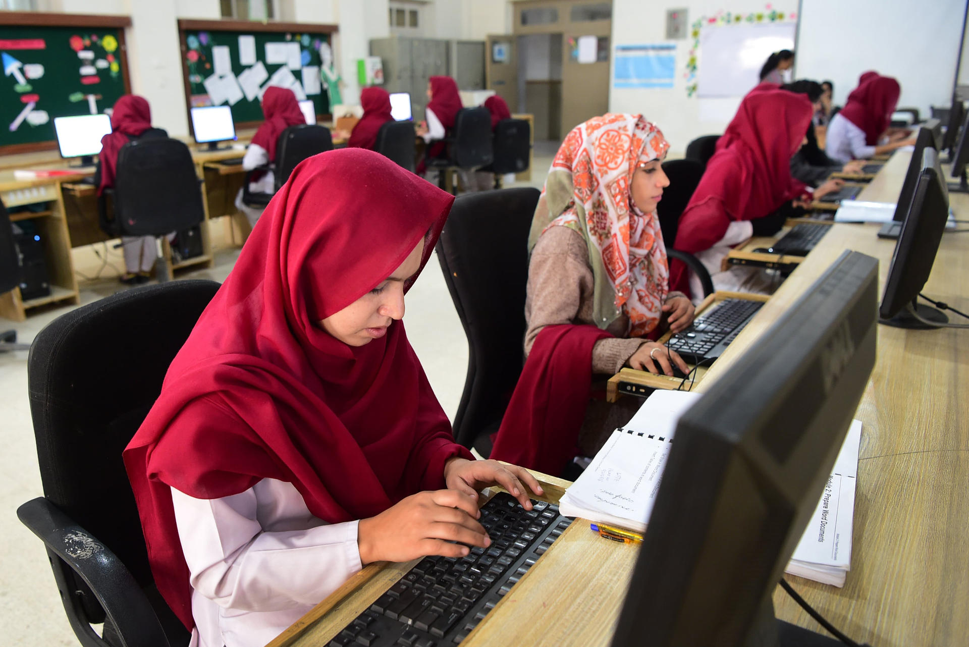 Student in red hijab typing on a desktop computer in a busy computer lab.