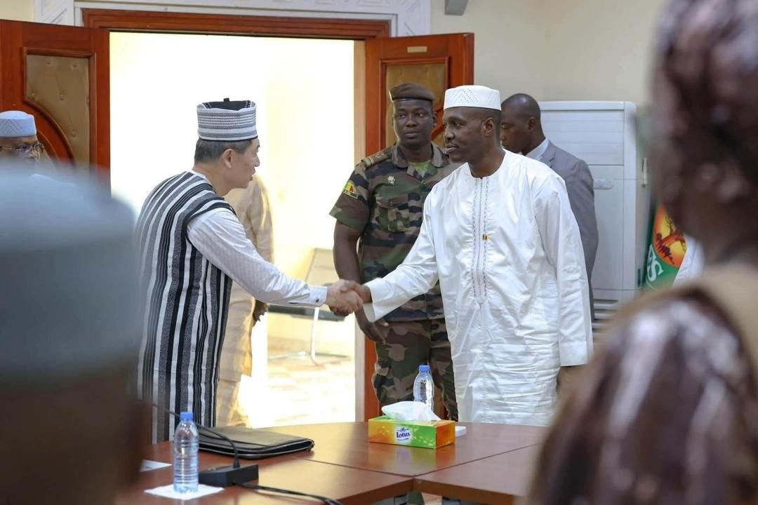 Photograph of two men in traditional attire shaking hands at a meeting; security in background.