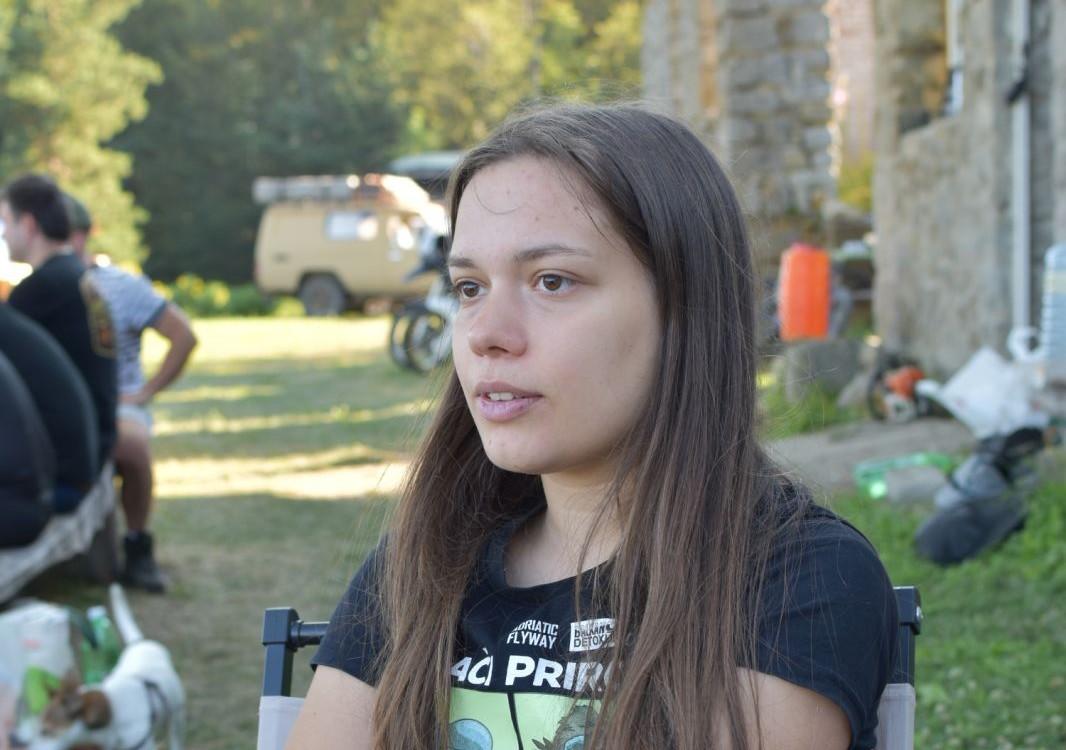 Photograph of a young woman with long brown hair outdoors, looking right, stone building behind.