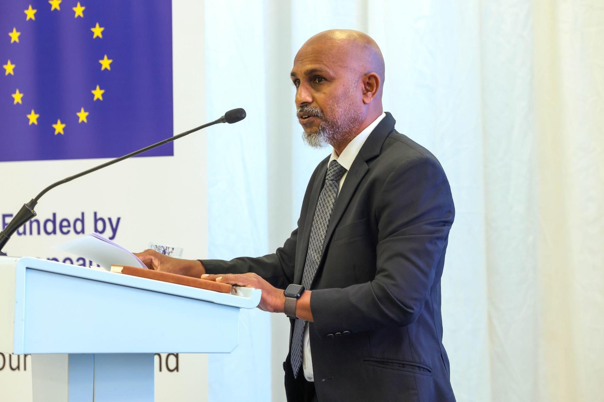 Man in a dark suit speaks at a podium with the European Union flag in the background.