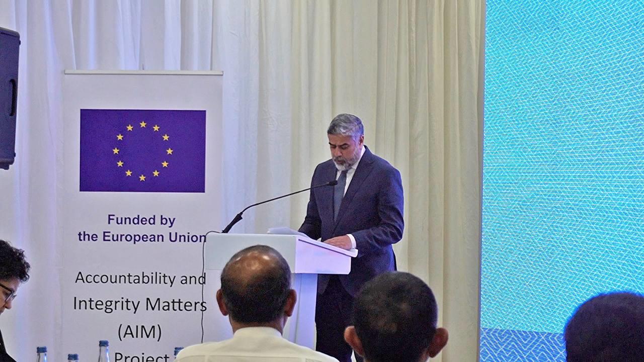 Male speaker at podium during conference; EU flag banner on left, blue screen on right.