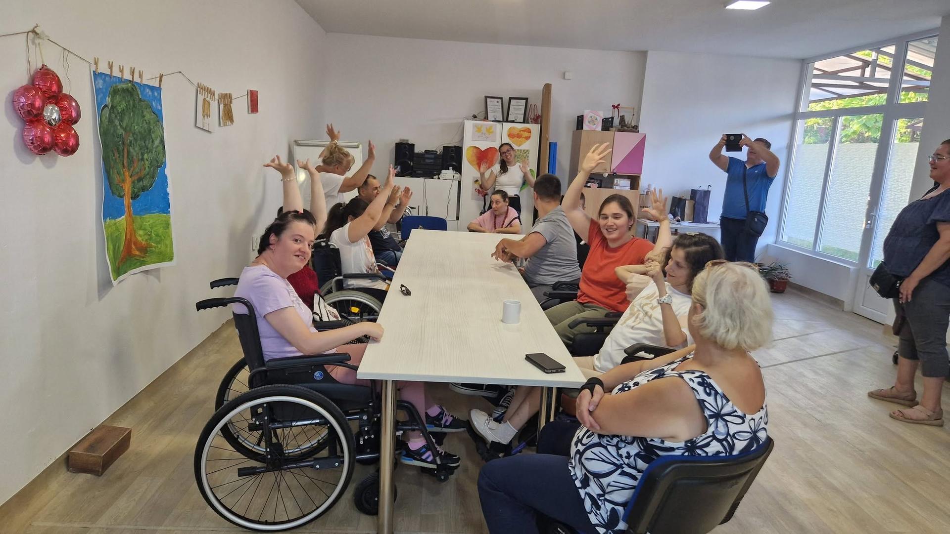 Elderly residents around a long table in a bright room with wall art; wheelchairs nearby.